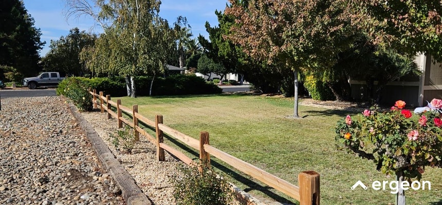 Wooden Cattle Fence in a Residential Yard in Reno This image shows a simple wooden cattle-style fence installed along the edge of a well-maintained residential lawn. The fence consists of vertical wooden posts with two horizontal wooden rails connecting them. The setting appears to be a suburban neighborhood with mature trees and bushes, including blooming rose bushes near the fence. The fence runs parallel to a gravel area, likely serving as a boundary marker to separate lawn space from a driveway or pathway. This type of fence is functional for marking property lines and providing a rustic aesthetic without fully obstructing views.