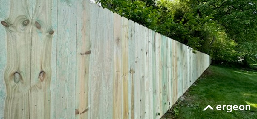 Long Wooden Privacy Fence in Richmond This image shows a long, newly installed wooden privacy fence stretching into the distance along a landscaped grassy area with trees in the background. The fence is constructed from vertical wooden planks with natural wood grain and knots visible, providing a sturdy barrier for privacy or boundary delineation. The lush greenery and mature trees behind the fence suggest it is in a residential or suburban outdoor setting. The image could be used in documentation concerning fence installation, local permit rules, or property boundary discussions in Richmond.