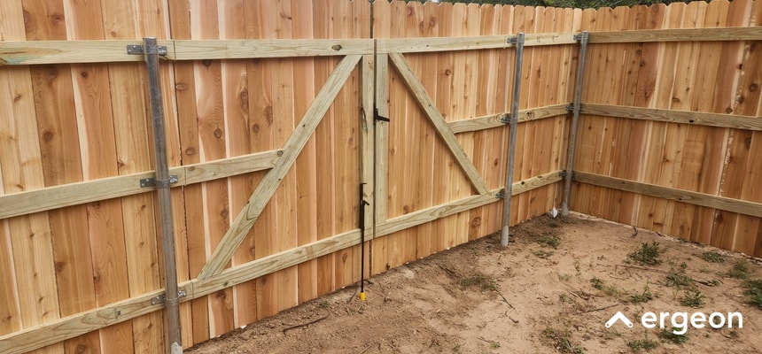 This image shows a corner section of a wooden privacy fence featuring vertical wooden planks and horizontal support beams, reinforced with metal brackets. The fence includes a double gate secured with metal hardware and supported by vertical metal posts embedded in the ground for stability. The area within the fenced space has a dirt surface with sparse vegetation, illustrating a typical residential outdoor enclosure suitable for property boundary definition and security.