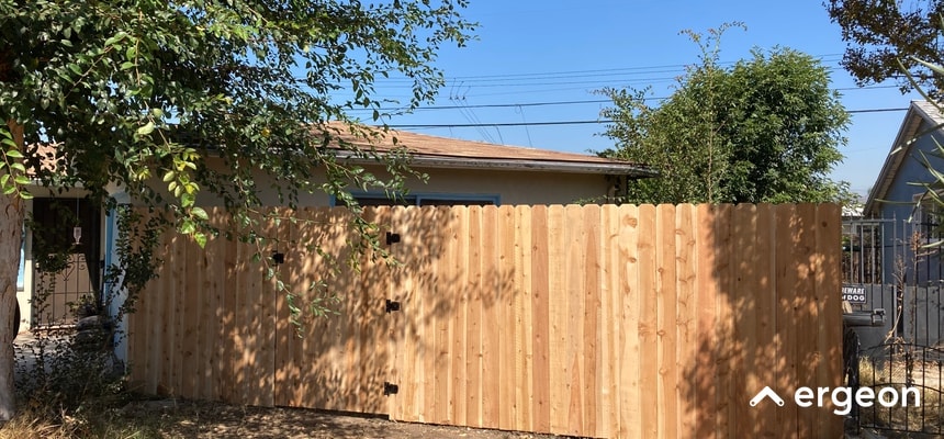 Wooden Privacy Fence for Residential Riverside Weather Protection This image shows a newly installed wooden privacy fence made from vertical wooden planks with rounded tops, secured with black metal hinges for gate access. The fence is located in a residential area with clear blue skies overhead. The surrounding trees suggest the fence is designed to withstand local riverside weather conditions, providing both privacy and security to the property it encloses.