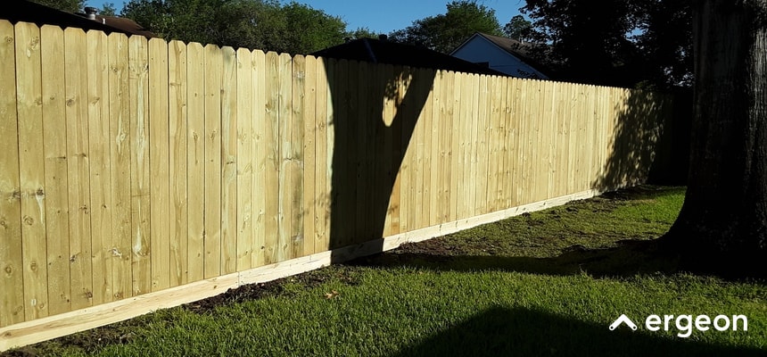 Wooden Privacy Fence Installed in a Residential Yard The image shows a long wooden privacy fence constructed with vertical wooden planks and a horizontal support board at the bottom. The fence appears newly installed, with fresh wood coloration and no visible weathering. It is situated in a residential backyard, bordered by grass and trees, and designed to provide privacy and security. The setting indicates it is well-suited for typical outdoor conditions, such as those found in Tampa weather, combining functionality with an aesthetic natural look.