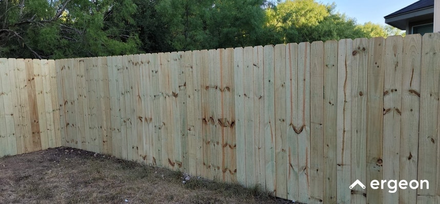 Wooden Privacy Fence Installation in Suburban Setting This image shows a newly installed wooden privacy fence in a suburban backyard. The fence consists of vertical wooden planks placed closely together to provide privacy and security. The wood appears untreated or lightly treated, with natural wood grain and knots visible. The fence is positioned on a flat patch of ground with some sparse grass and small plants at the base. In the background, there are trees and a portion of a house roof visible, indicating a residential setting. This type of fence is commonly used to define property boundaries and enhance privacy while maintaining a natural aesthetic.