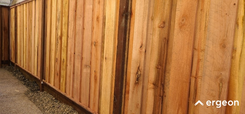 Wooden HOA-Compliant Fence in Watsonville This image depicts a professionally constructed wooden fence made from vertical cedar planks with natural wood variations, installed along a gravel and concrete pathway. The fence demonstrates typical Homeowners Association (HOA) compliance for residential properties in Watsonville, featuring clean, straight lines and sturdy posts, reflecting quality craftsmanship and adherence to local neighborhood guidelines. The image includes the 'ergeon' logo, indicating the source or related service provider.