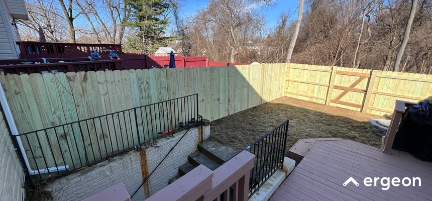 New Wooden Fence Installation in Residential Backyard in Wilmington This image shows a recently installed wooden privacy fence enclosing a residential backyard in Wilmington. The fence is constructed using light-colored vertical wooden planks and includes a matching gate. The backyard also features a patio deck area with wooden flooring and metal railings leading down to a lower level with concrete steps. The area appears well-maintained with a grass lawn and leafless trees indicating a winter or early spring season. The image is suitable for illustrating fence permit rules, construction standards, or residential property boundary guidelines.