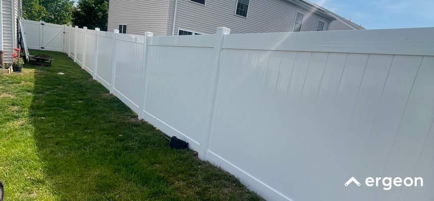 White Vinyl Privacy Fence Installed in Residential Backyard The image shows a long stretch of a white vinyl privacy fence installed along the boundary of a residential backyard. The fence panels are solid, providing complete privacy, and are supported by evenly spaced vertical posts with decorative post caps. The fence runs parallel to a house wall on a well-maintained grassy lawn, under clear daylight conditions. The image highlights the clean, modern design and durable material of the fence, suitable for knowledge sharing in home improvement, landscaping, and fencing regulation contexts.