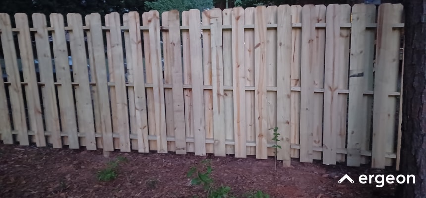 Wooden Privacy Fence Built for Tallahassee Weather Conditions This image shows a recently installed wooden privacy fence constructed from vertically aligned wooden planks with a protective finish. The fence is designed to withstand Tallahassee's weather, featuring durable treated wood panels and horizontal support beams for added stability. The fence is set on bare soil with some sparse plant growth visible, and the backdrop includes dense trees. This type of fence provides privacy and security while being suited for local climate conditions.