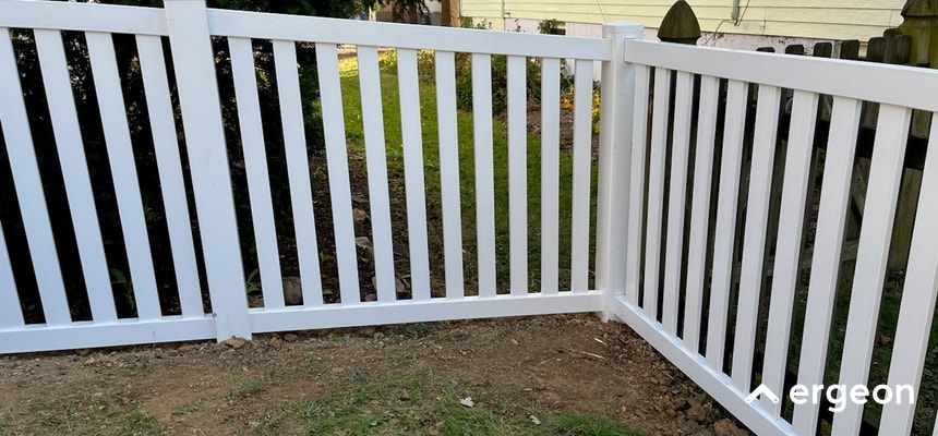White Vinyl Fence Corner Installation in Virginia Beach This image shows a newly installed white vinyl fence featuring a clean corner joint with vertical slats and horizontal supports. The fence is set up on a patch of bare soil, with a background of grass, shrubs, and a wooden fence. The high-quality vinyl material and secure corner connection suggest durability and aesthetic appeal, making this fence suitable for residential boundary definition or garden enclosures in Virginia Beach or similar locations.