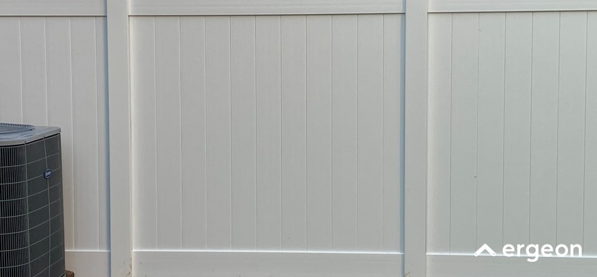 White Vinyl Fence Installed Adjacent to Outdoor Air Conditioning Unit This image shows a clean, white vinyl fence with vertical paneling. On the left side of the fence, there is a visible outdoor air conditioning unit. The fence is constructed with sturdy posts and panels, providing privacy and a modern aesthetic. The photo highlights the fence's installation quality and its integration into a residential or commercial property environment. The branding 'ergeon' is visible in the lower right corner of the image, indicating the company associated with the fencing solution.