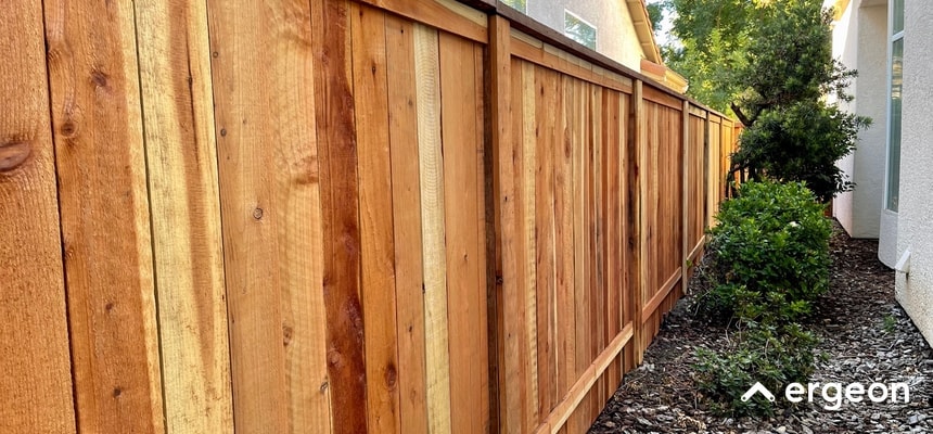 Wooden Privacy Fence Installed Between Residential Homes This image shows a newly installed wooden privacy fence running along the side of a residential property. The fence is constructed from vertical wooden planks with horizontal support beams, providing a secure and aesthetically pleasing boundary. To the right of the fence, there is a narrow landscaped area with soil, mulch, and several green shrubs. The scene highlights typical fencing used for privacy and boundary definition in suburban areas. The image can be used for reference in residential fencing design, installation guides, or property boundary discussions.
