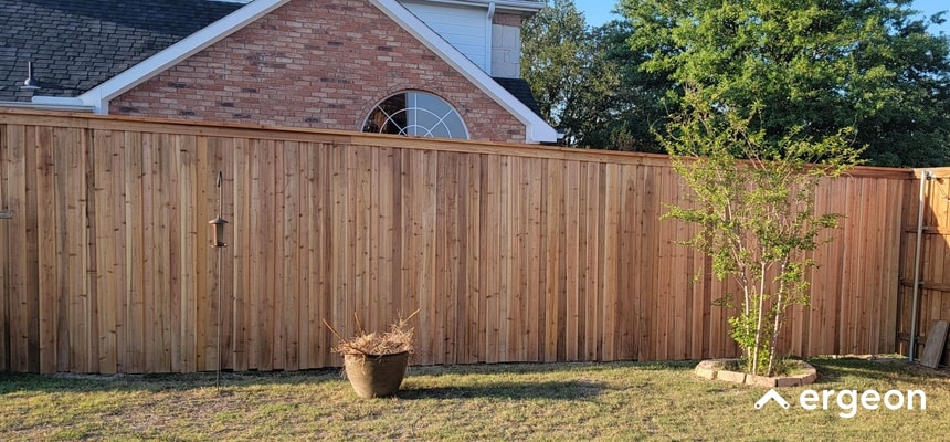 Wooden Privacy Fence Installed in Residential Backyard This image displays a newly installed wooden privacy fence enclosing a residential backyard. The fence is constructed from vertical wooden planks with a natural finish, providing a solid barrier for privacy and security. In the yard, there is a potted plant with dried foliage and a small tree planted in a circular mulched bed near the fence. The setting includes a grass lawn, a brick house with a triangular roofline, and some trees in the background. This visual can be used to illustrate examples of privacy fencing options and backyard landscaping.