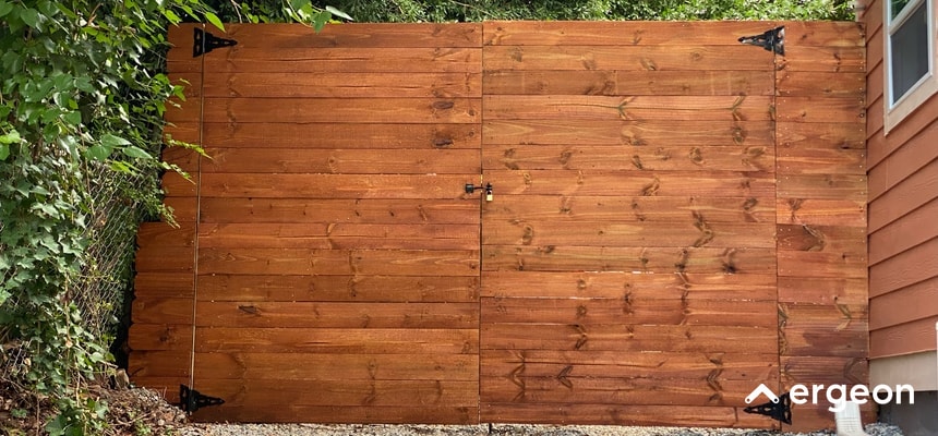 Wooden Fence Gate Featuring Sturdy Hinges and Lock This image depicts a wooden fence gate constructed from horizontal wooden planks with a smooth finish. The gate includes visible black metal hardware such as robust hinges on the left and right edges and a central locking latch secured with a padlock. The gate is installed between a dense foliage area on the left and an exterior wall of a house on the right, providing a secure and natural-looking barrier. This image is suitable for use in documentation or guides related to fence construction, gate security, or outdoor home improvement projects.