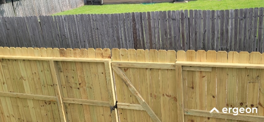 This image shows a recently installed wooden fence in the foreground with fresh, light-colored wood panels and a gate equipped with a latch. Behind it is an older, weathered wooden fence with darker, grayish panels, indicating exposure to the elements over time. The photo highlights the contrast between new construction and aged fencing, useful for illustrating fence replacement or upgrade projects.