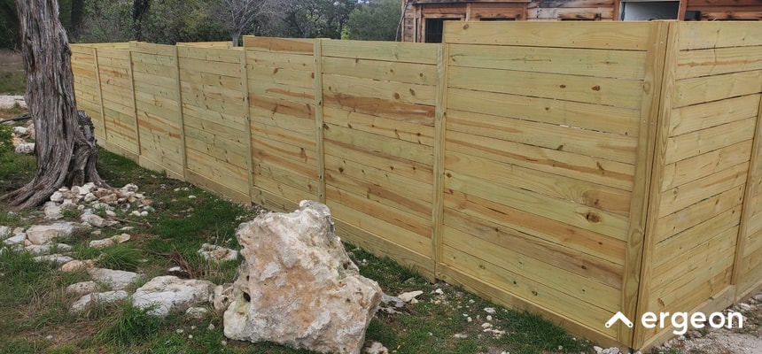 Wooden Privacy Fence Installed in Outdoor Yard The image shows a newly installed wooden privacy fence extending along a property boundary in an outdoor yard. The fence is constructed from horizontal wooden planks supported by vertical posts at regular intervals. The wood is light-colored, likely untreated or freshly installed, and the fence provides a solid barrier for privacy. The ground around the fence is natural earth with grass, rocks, and a tree visible, indicating a rural or suburban setting. This type of fence is typically used for securing and defining residential or private outdoor spaces.
