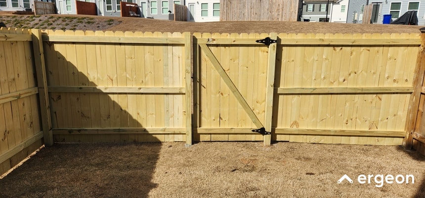 Wooden Fence with Gate in Residential Backyard This image shows a newly installed wooden privacy fence in a residential backyard. The fence is constructed using vertical wooden planks and includes a central gate secured with black metal hinges and latch hardware. The fence encloses the backyard area, which has dry grass, and neighboring houses are visible beyond the fence. The image demonstrates a typical example of a wood fence installation used for privacy and boundary demarcation in suburban homes.