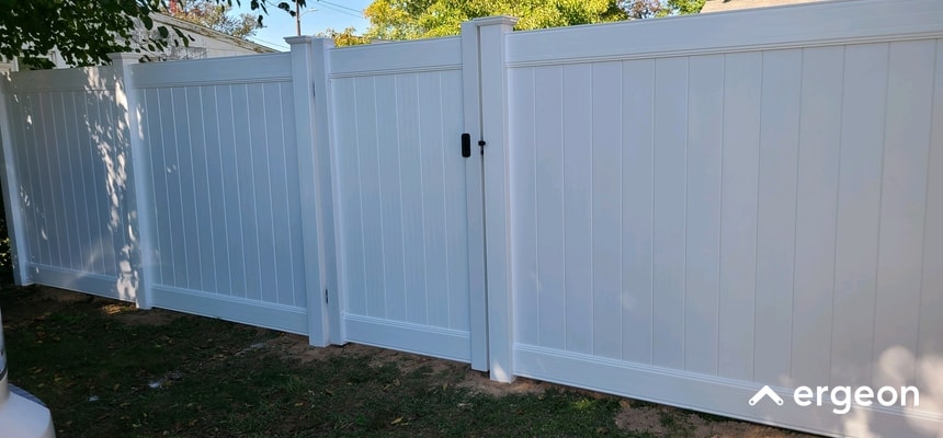 White Vinyl Privacy Fence with Gate and Lock This image shows a section of a white vinyl privacy fence featuring vertical panels and posts. The fence includes a central gate equipped with a black locking mechanism and handle, demonstrating the security features typical of residential outdoor fencing. The scene is taken in daylight with some trees casting shadows, illustrating the fence's aesthetic and functional purpose in providing privacy and boundary delineation for a yard or property.
