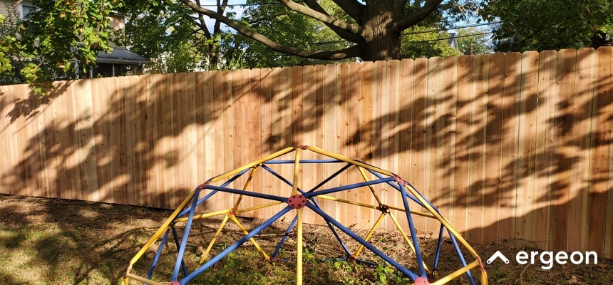 New Wooden Fence Installed in Backyard with Playground Climbing Dome The image shows a newly installed wooden privacy fence running along the edge of a backyard. The fence is made of vertical wooden planks with a scalloped top edge. In front of the fence, there is a children's playground climbing dome structure, constructed with blue and yellow metal bars. The background includes tree foliage and residential buildings partially visible through the trees. This setup illustrates a fenced backyard area that provides privacy and safety, with recreational equipment for children.