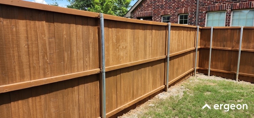 This image shows a close-up view of a wooden privacy fence meeting at a corner, reinforced with vertical metal support posts. The fence consists of vertical wooden boards secured with horizontal wooden rails, providing sturdy construction. The metal posts offer additional structural support, ensuring stability and durability. The fenced area encloses a grassy yard adjacent to a brick residential building. This image is useful for demonstrating fence construction techniques relevant to site planning and permit applications.