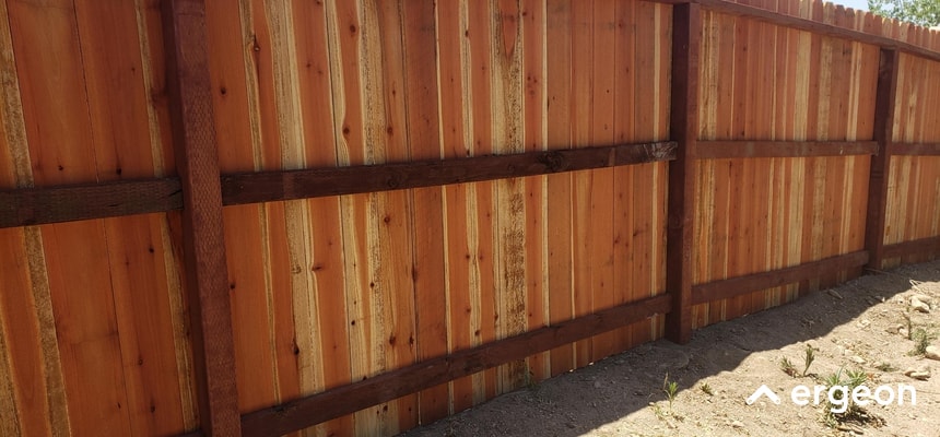 This image shows a newly installed wooden privacy fence panel made of vertical wooden planks supported by horizontal and vertical support beams. The fence is set up outdoors on bare soil with sparse vegetation, providing privacy and boundary definition for a property. The lighting indicates a sunny day, highlighting the natural color and texture of the wood. This type of fence is commonly used for residential boundary security and enhancing backyard privacy.