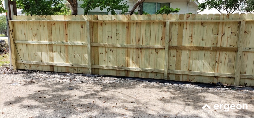 This image shows a newly constructed wooden privacy fence in a residential outdoor setting. The fence is made of vertical wooden planks secured by horizontal support beams and evenly spaced posts. The wood appears fresh and untreated, indicating recent installation. The fence is designed to provide privacy and boundary definition for the property visible in the background, including a house window partially seen behind the fence. The ground in front of the fence is a mix of dirt and small rocks. This type of fence installation is typically used for enhancing security, privacy, and aesthetic appeal in residential areas.