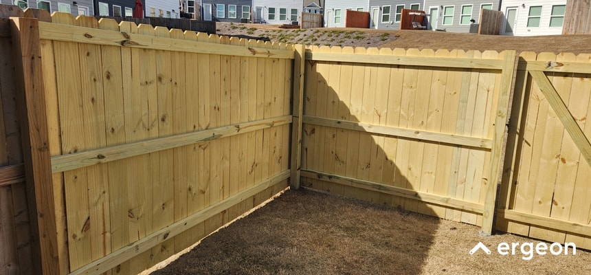 This image displays a newly installed wooden fence corner in a residential backyard. The fence panels are made of vertical wooden planks supported by horizontal wooden rails and sturdy corner posts. The ground shows dry grass typical of a backyard setting. In the background, neighboring houses and additional fencing are visible. This image can be used for illustrating fence construction, residential fencing options, or landscaping boundaries in professional home improvement or landscaping documentation.