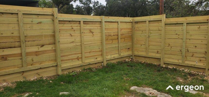 This image shows a recently installed wooden fence forming a corner enclosure. The fence consists of vertical posts supporting horizontal wooden panels with a natural light wood finish. The fence is constructed on a grassy outdoor area with some rocks on the ground near the base. The design appears sturdy and uniform, suitable for residential or commercial property boundary demarcation and privacy. The fence structure emphasizes craftsmanship and material quality, providing a clear example for professionals or customers seeking fencing solutions.