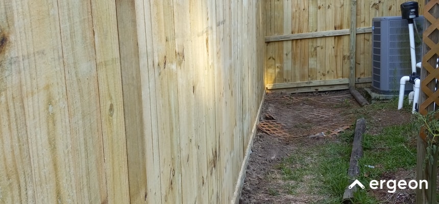 This image shows a newly installed wooden privacy fence running alongside a residential property. The fence panels are vertically aligned and made of untreated natural wood. In the background, there is an HVAC outdoor unit and some plumbing components. The ground beside the fence is partly covered with grass and dirt, indicating recent landscaping or construction activity. The photo illustrates a typical setup where fencing is installed close to home utility equipment, highlighting space management and outdoor structural work.