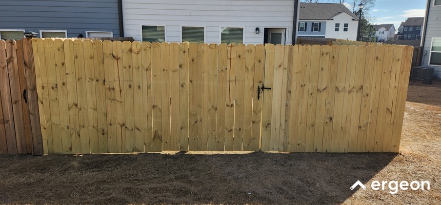 The image shows a newly installed wooden privacy fence with a double gate in a residential backyard. The fence is constructed from natural, unfinished wooden vertical planks, providing full privacy. The gate features a central black metal latch and hinges, indicating it is functional and secure. The fence is situated on dry soil with a row of modern houses visible in the background. This image is useful for illustrating fence installation types, material use in residential fencing, and gate design considerations.
