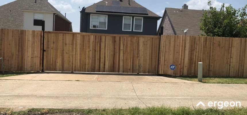 This image shows a tall wooden privacy fence enclosing a residential backyard with a double wooden gate providing access from the driveway. The fence is made of vertical wooden planks with a gate latch mechanism visible on the left side. In the background, there are neighboring houses with pitched roofs, and the foreground shows a concrete driveway leading up to the closed gate. The image emphasizes secure boundary definition around the property, suggesting privacy and security considerations. A small ADT security sign is displayed near the gate, indicating a monitored alarm system. The branding 'ergeon' appears in the bottom right corner.