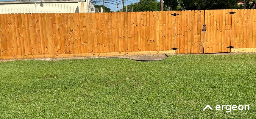 This image depicts a residential backyard with a neatly maintained green lawn in the foreground and a tall wooden fence running across the background. The fence includes a gate secured with black hardware. The area appears to be well-kept, illustrating a typical private outdoor space that might be referenced in discussions relating to site planning, landscaping, fencing, or residential property management.