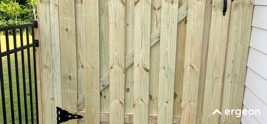 This image shows a close-up view of a new wooden gate constructed with vertical wooden planks reinforced by diagonal wooden supports on the backside. The gate is secured with a black metal hinge at the bottom left corner. To the left of the wooden gate is a black metal fence with vertical bars. Part of a light-colored building wall is visible on the right side of the image. The ground beneath the gate is visible, showing the connection between the gate and the rest of the fence. The surrounding background includes green grass and trees, suggesting an outdoor setting. This type of gate and fence can be relevant for site plans or permit applications related to residential property boundaries and access control.