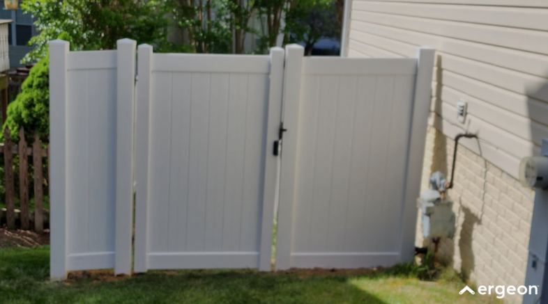 This image shows a white vinyl privacy gate attached to a matching vinyl fence. The gate features vertical panels with a central locking mechanism and is set on grass adjacent to a beige house exterior wall. Utility meters and pipes are visible mounted on the house near the gate, indicating the gate provides access between the front and backyard areas. The setup is typical for residential properties seeking a durable, low-maintenance fencing solution for privacy and security.