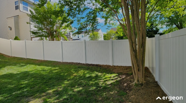 The image shows a long white vinyl privacy fence bordering a residential backyard. The fence panels are vertically lined and connected with sturdy posts, creating a clean and uniform appearance. The backyard area features a grassy lawn with a tree near the corner where the fence turns at a right angle. In the background, there are neighboring houses and additional trees under a partly cloudy sky. This type of fencing is typically used for residential properties to provide privacy, security, and aesthetic appeal.