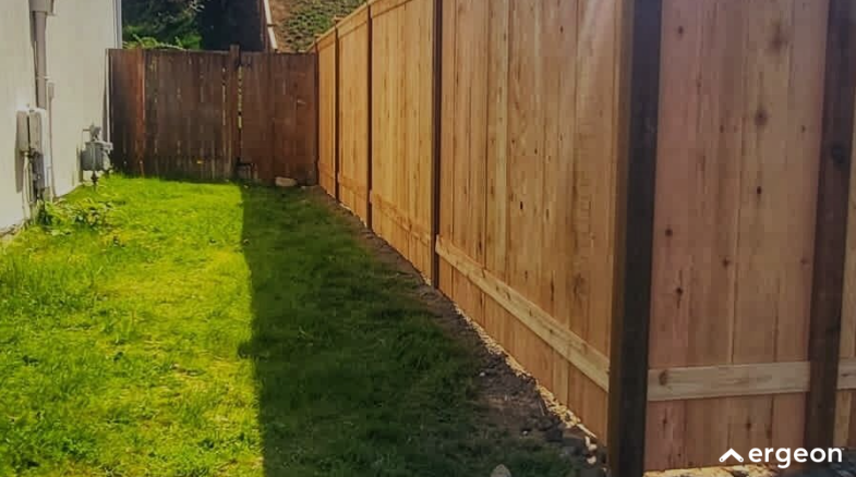 This image shows a narrow residential backyard area featuring a wooden privacy fence on the right side and a white exterior wall of a house on the left. The ground is covered with green grass, with a shadow cast over part of the lawn. The fence appears recently installed or well-maintained, and there is a wooden gate at the far end of the yard. The setting suggests a private outdoor space typical of suburban homes, useful for landscaping, fencing design, or property boundary documentation.
