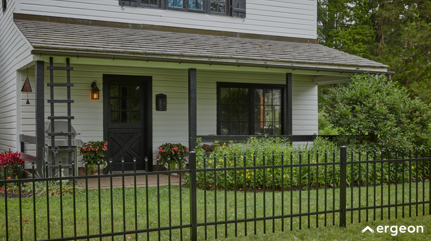This image shows the front porch of a white cottage-style house featuring a prominent black door with window panes and a multi-paned front window. The porch has a roof supported by black posts and is decorated with lush green plants and flowers in pots. A black metal fence with vertical bars encloses the green front lawn, adding a layer of security and aesthetic appeal. The porch area includes a small step up from the walkway and a decorative element resembling a wooden trellis attached to the side. The setting is serene with surrounding greenery, suggesting a cozy and well-maintained residential property. This image can be used in real estate, architectural design, or home landscaping documentation.