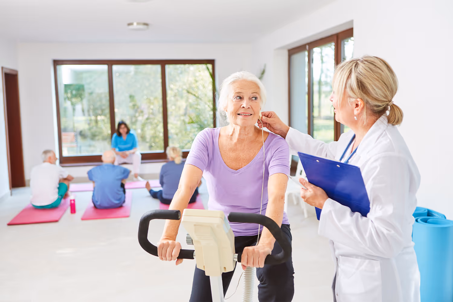 Elderly woman riding a stationary bicycle with an exercise physiologist monitoring her.