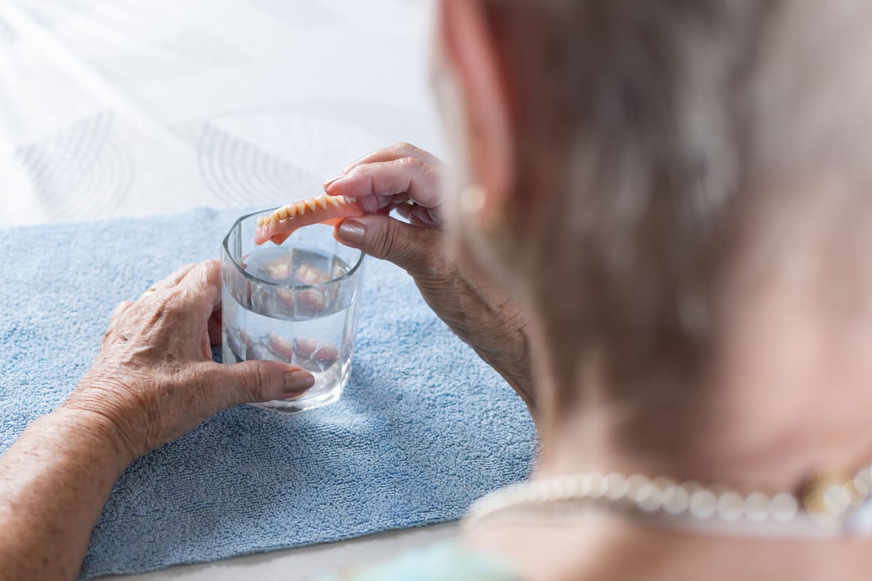 Elderly woman placing dentures into glass of water for proper overnight storage and cleaning