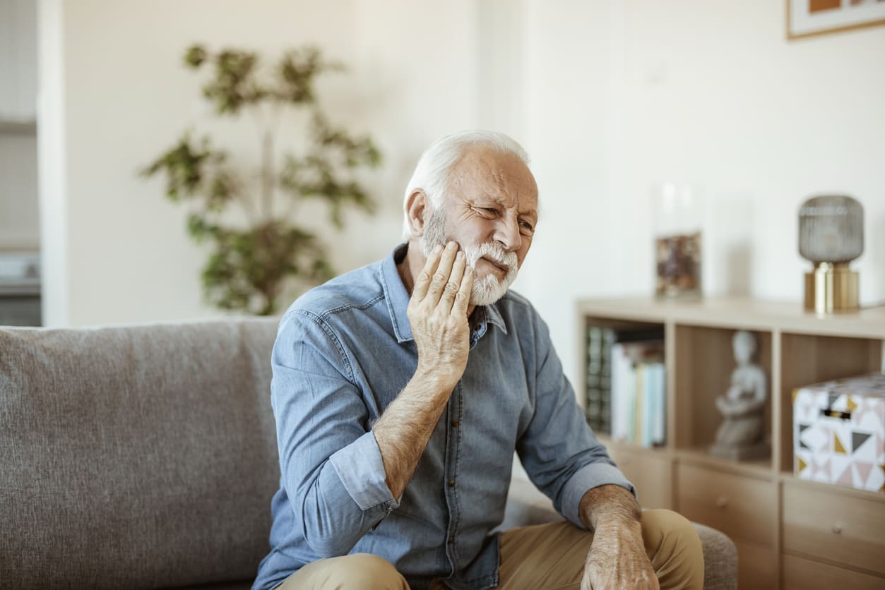 Senior man sitting on couch holding jaw in pain from denture sore spots causing eating discomfort
