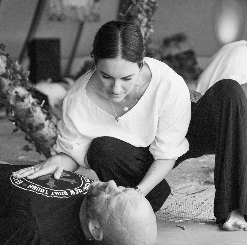 Aliya kneeling beside a man lying on a mat, one hand resting on his chest, holding space for him