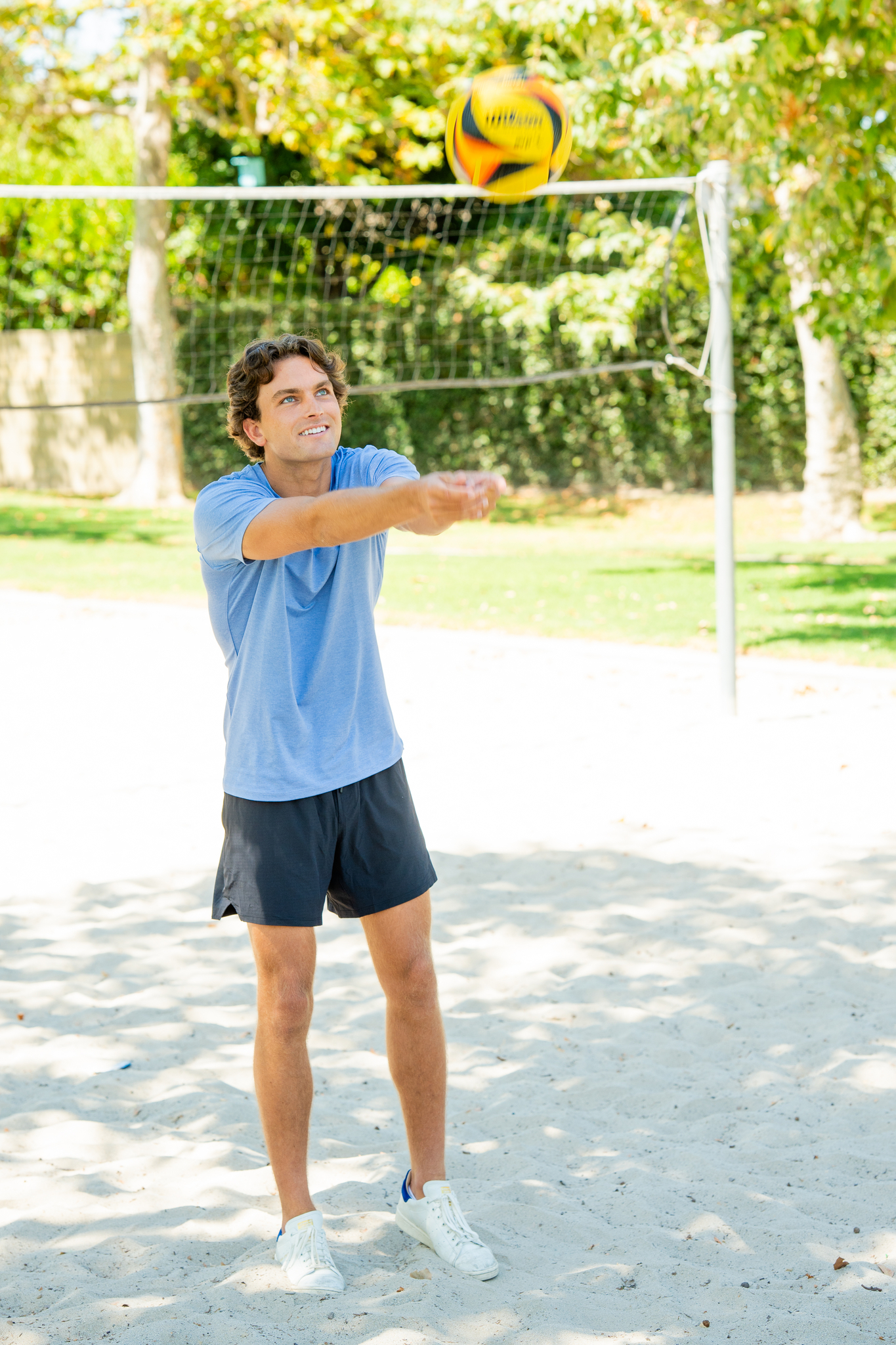 Young man in a light blue t-shirt and black shorts playing volleyball on a sandy outdoor court, hitting a yellow and black volleyball with his forearms.