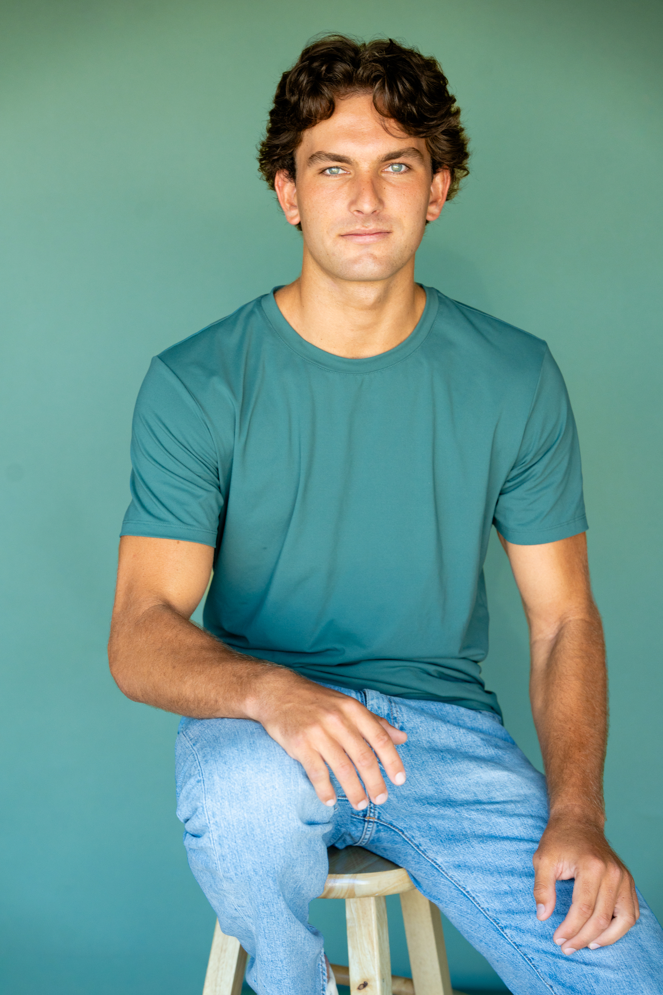 Young man with curly brown hair and blue eyes wearing a teal t-shirt and light blue jeans sitting on a wooden stool against a teal background.