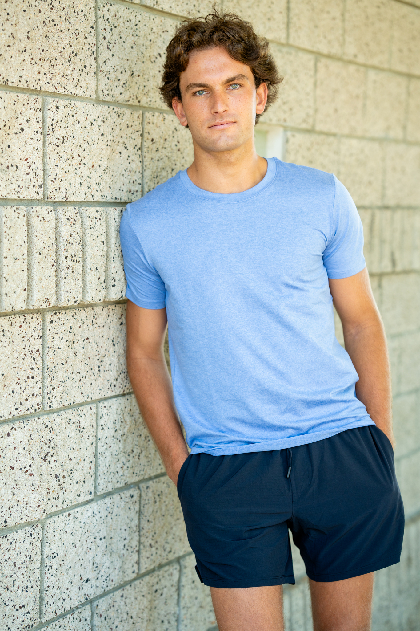 Young man with curly brown hair and blue eyes wearing a light blue t-shirt and dark shorts, standing with hands in pockets and leaning against a light stone brick wall.