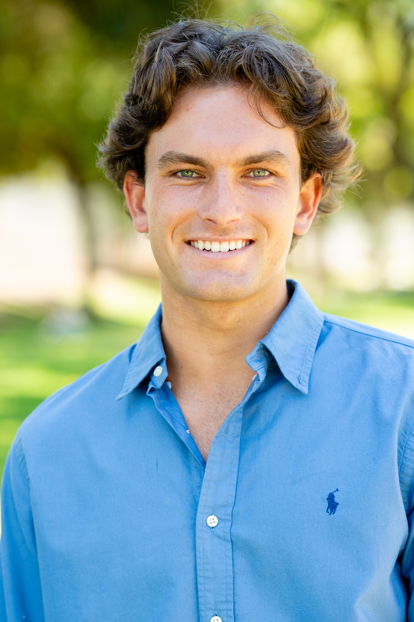 Portrait of a young man with wavy brown hair and green eyes, smiling and wearing a blue button-up shirt outdoors with a blurred green and yellow background.