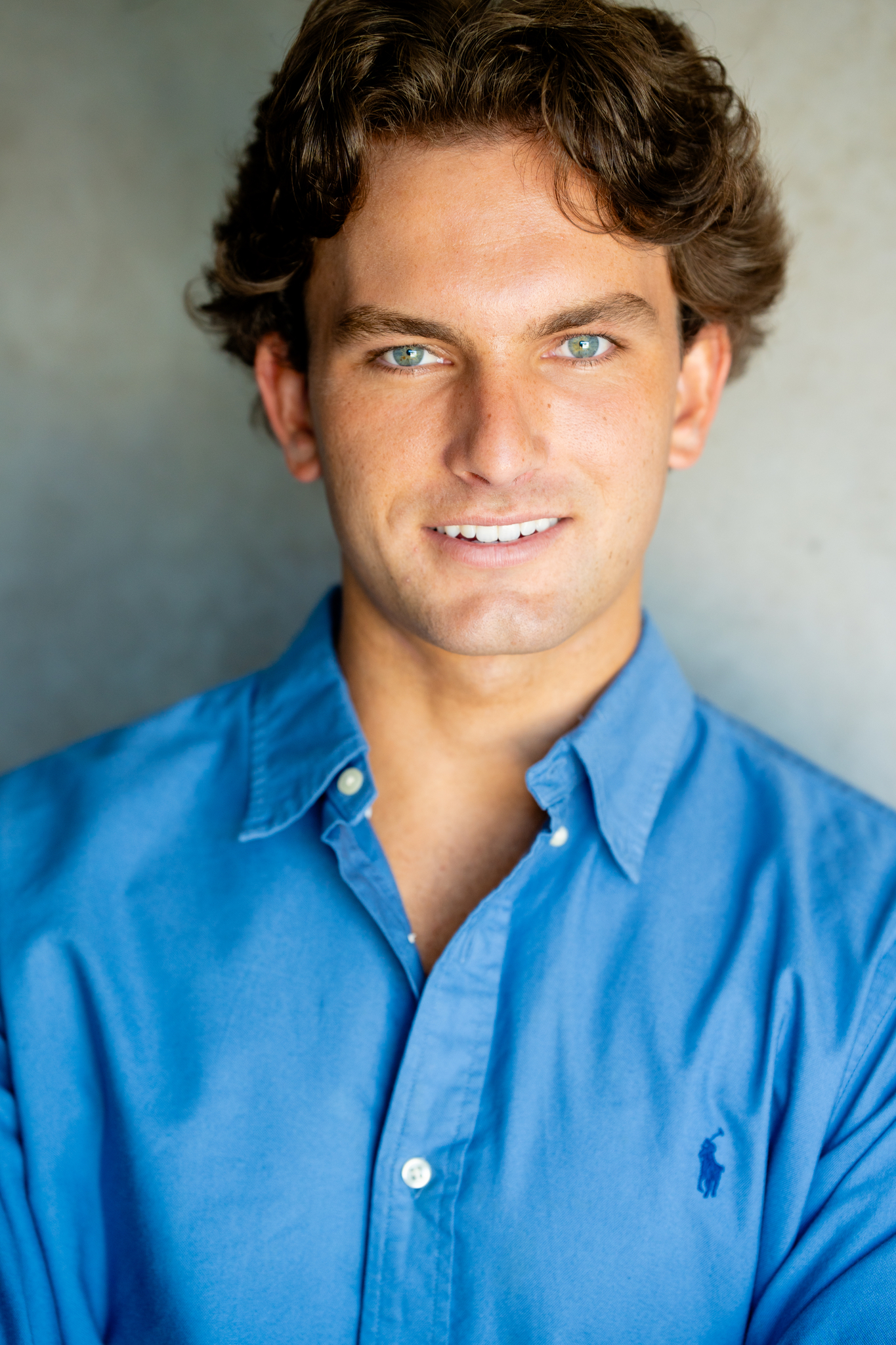 Portrait of a young man with blue eyes and curly brown hair wearing a blue button-up shirt with a small polo logo, smiling slightly against a plain gray background.