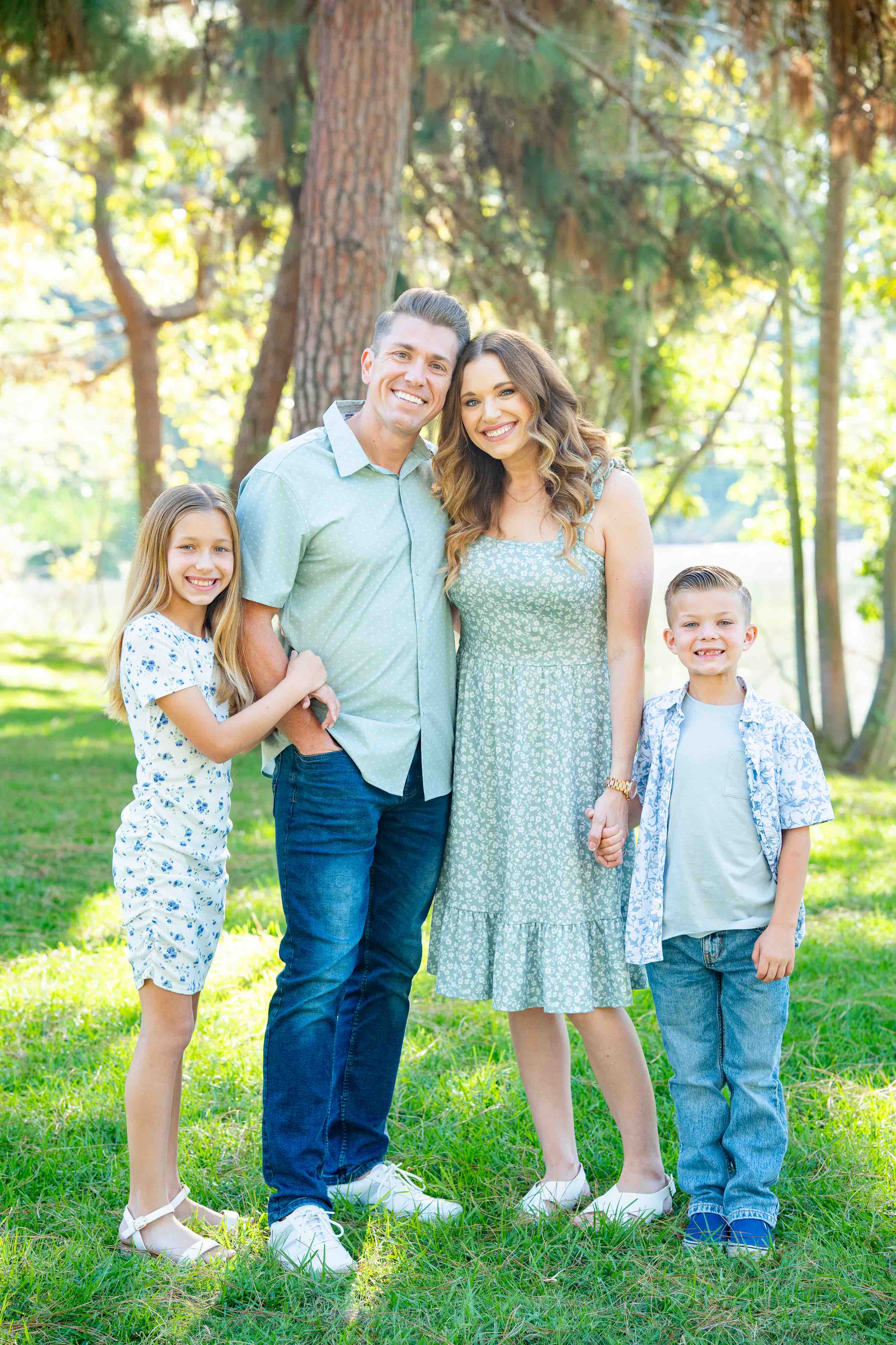 Smiling family of four standing on grass in a park with trees in the background, including a man and woman in green clothing, and two children with light hair wearing casual outfits.