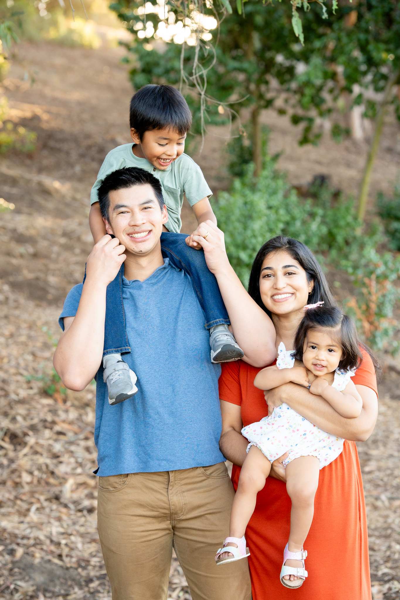 Smiling family of four outdoors with father wearing a blue shirt and tan pants carrying young boy on his shoulders, and mother in an orange dress holding a toddler girl in a white floral outfit.