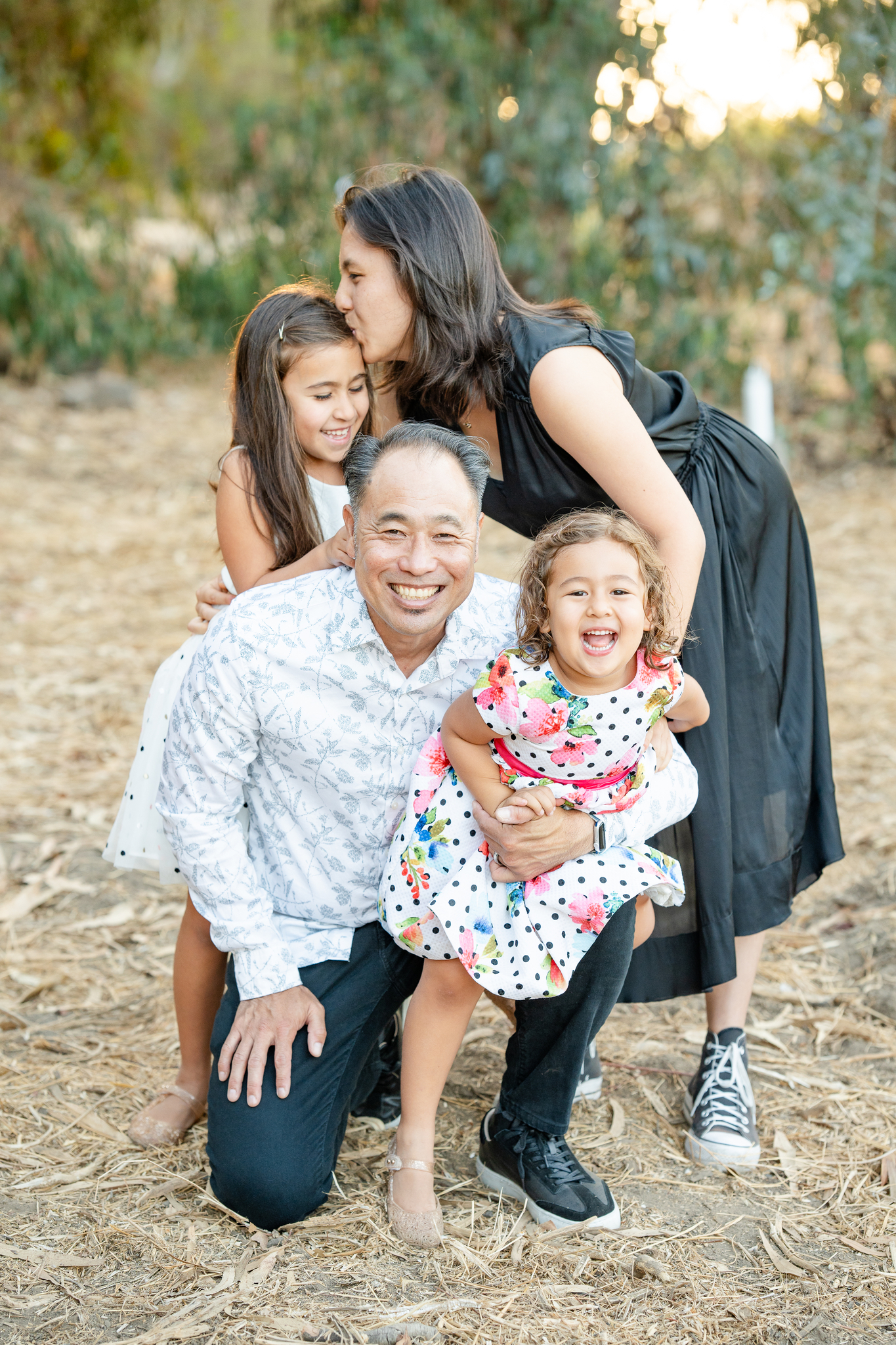 An adult man kneeling outdoors smiles while holding a young girl in a floral dress and polka dots, who is laughing. Behind him, an adult woman in a black dress kisses the forehead of an older girl in a white dress with black dots who is smiling and leaning on the man's shoulder. The background shows trees and soft sunlight.