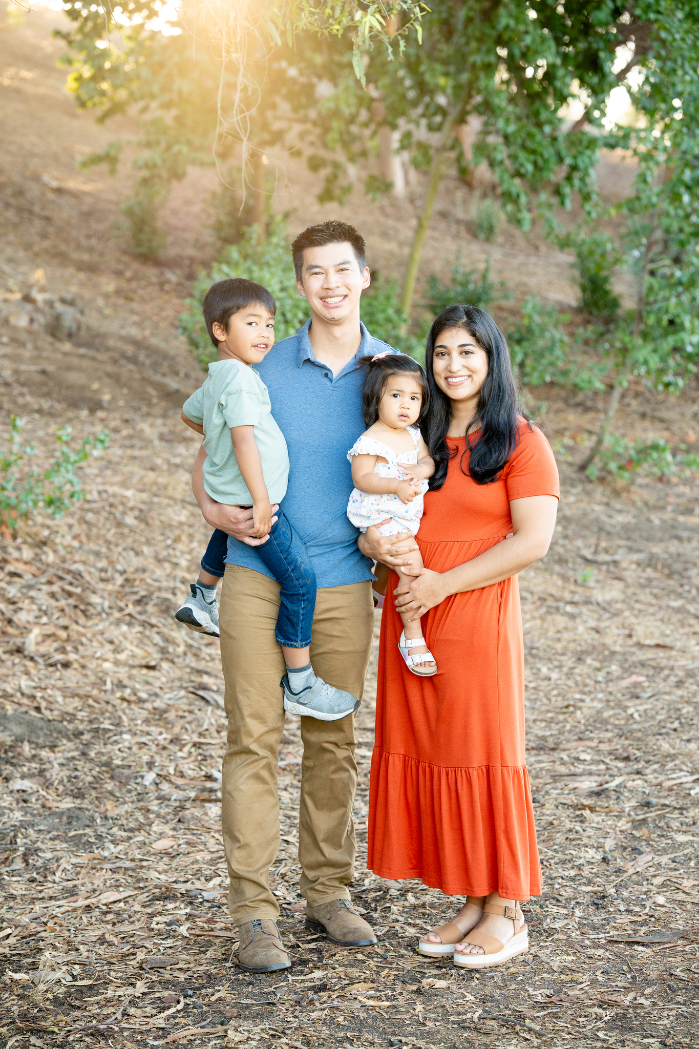 Smiling family of four standing outdoors on dry leaf-covered ground with green trees in the background, the father in a blue polo and tan pants holding a young boy, the mother in an orange dress holding a toddler girl in a white dress.
