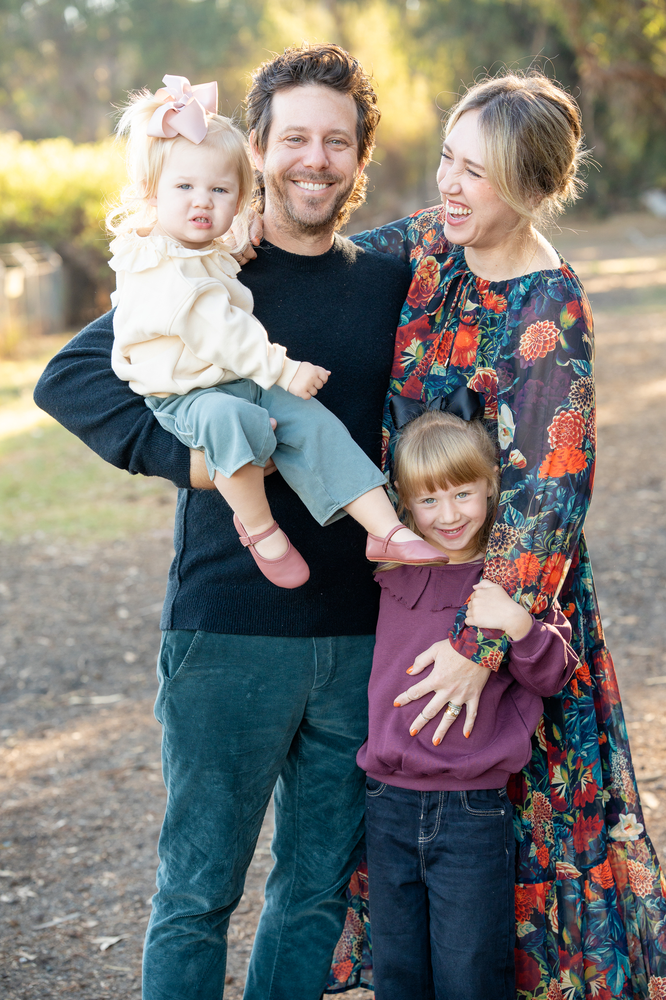 A smiling man holds a toddler with a pink bow in her hair and pink shoes, while a laughing woman in a floral dress embraces a smiling girl with a black bow and purple sweater outdoors.