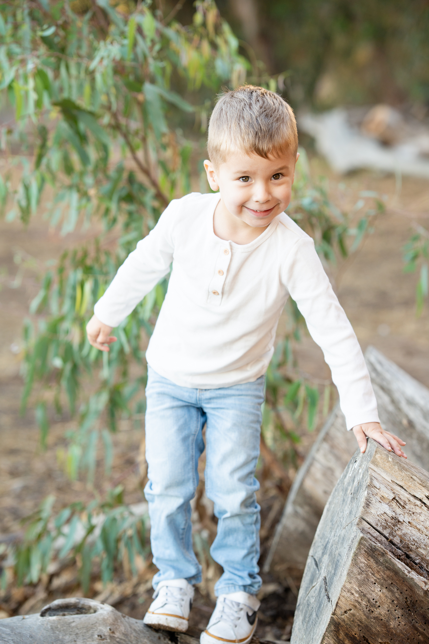 Young boy wearing a white long-sleeve shirt and light blue jeans balancing on tree logs outdoors, smiling at the camera with green leaves in the background.