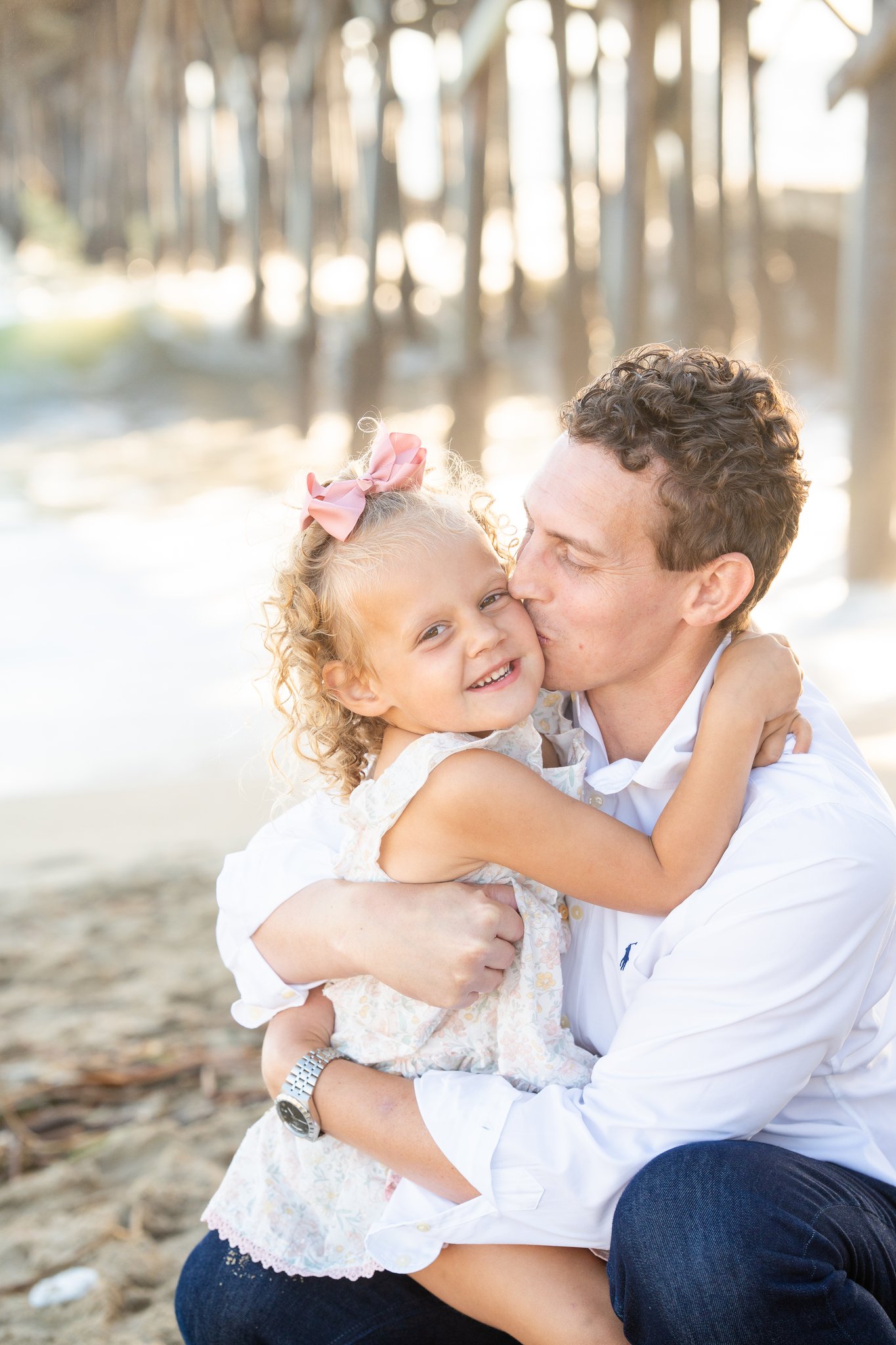 A man in a white shirt sits on sand under a pier, hugging and kissing a smiling young girl with curly hair and a pink bow in her hair.